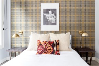 Eclectic bedroom with plaid wallpaper, matching brass sconces, wood nightstands, black and white photo, southwestern pillow.
